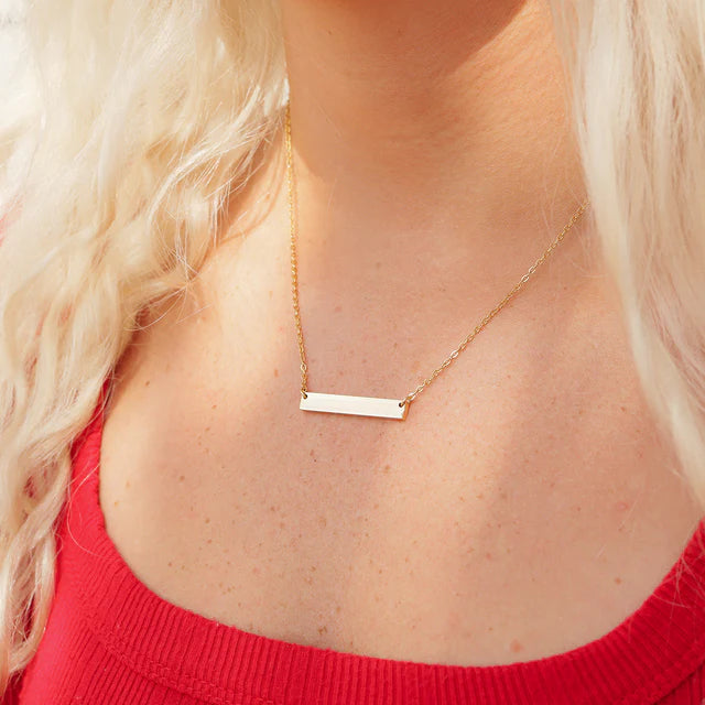 Gold bar necklace worn by a person with blonde hair and a red top.