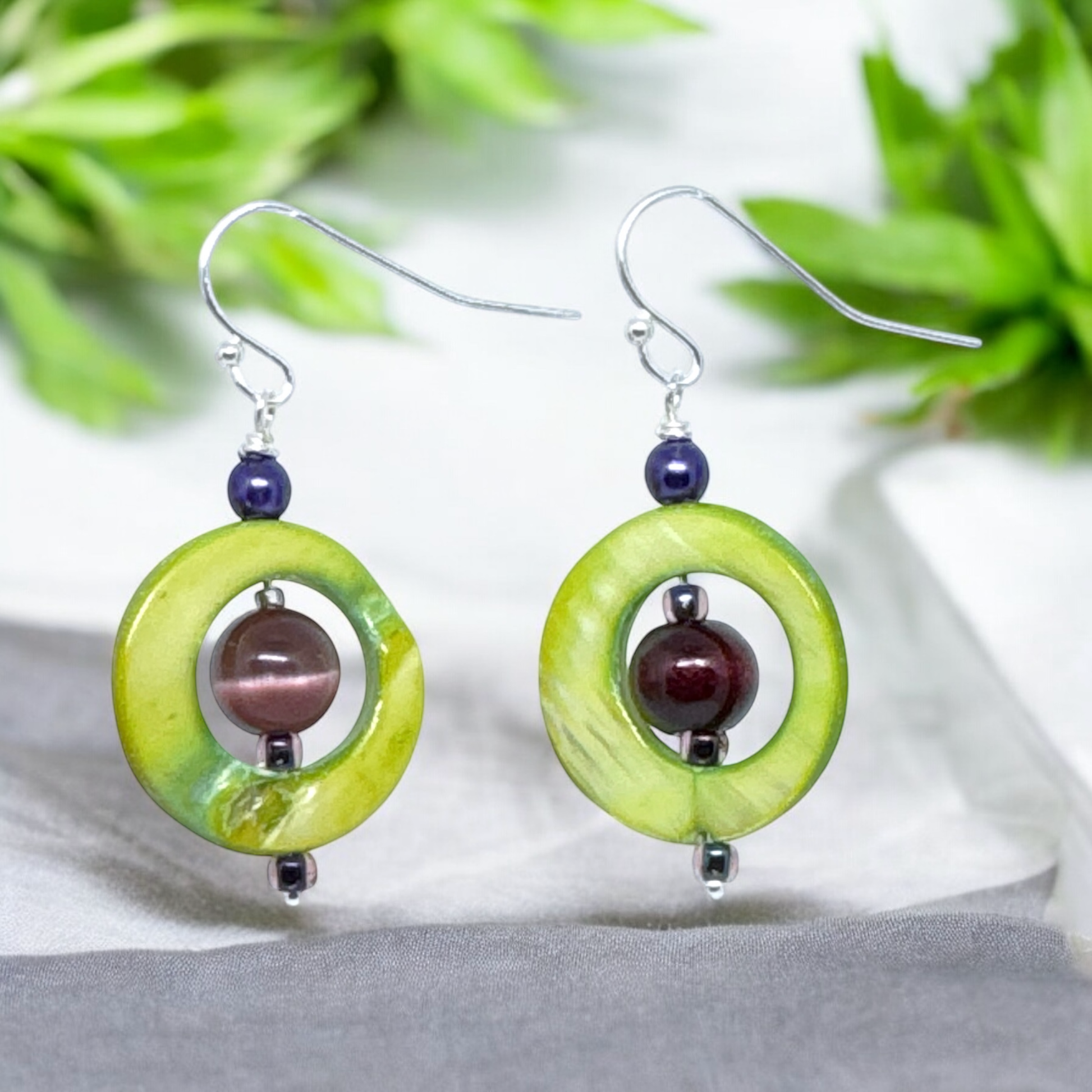 Close-up view of green shell and amethyst dangle earrings featuring circular shell pieces with natural variations and a purple amethyst bead in the center, showcasing their unique design.