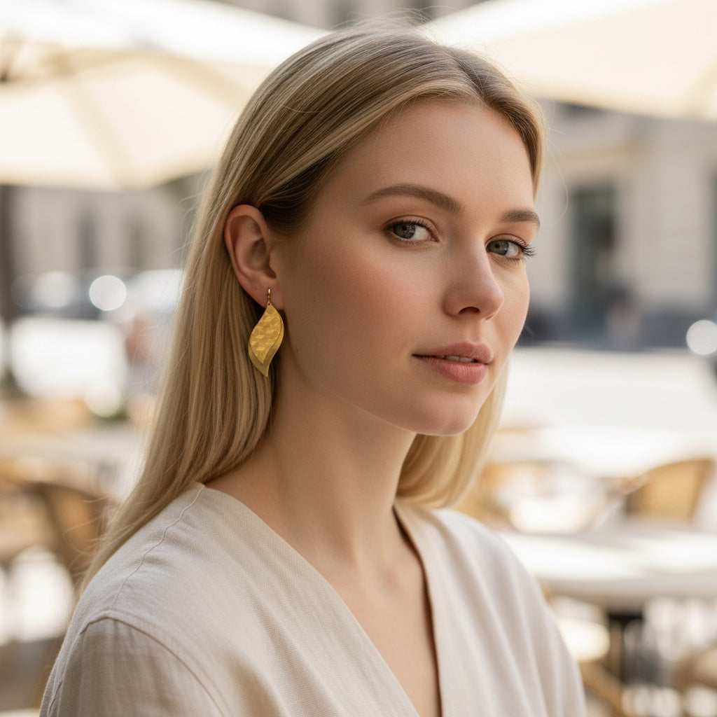 Bedrock Rose gold leaf dangle earrings with geometric etched brass layers worn by a blonde woman at an outdoor cafe.