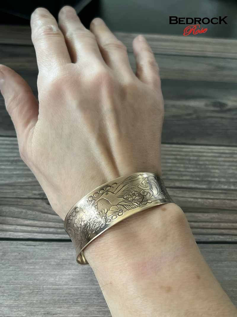 Bedrock Rose polished brass cuff bracelet featuring an intricate engraved hummingbird and floral design on a wrist.