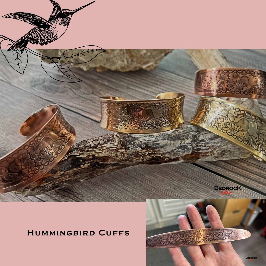 Bedrock Rose hummingbird cuffs in polished copper and brass with floral engravings displayed on driftwood.