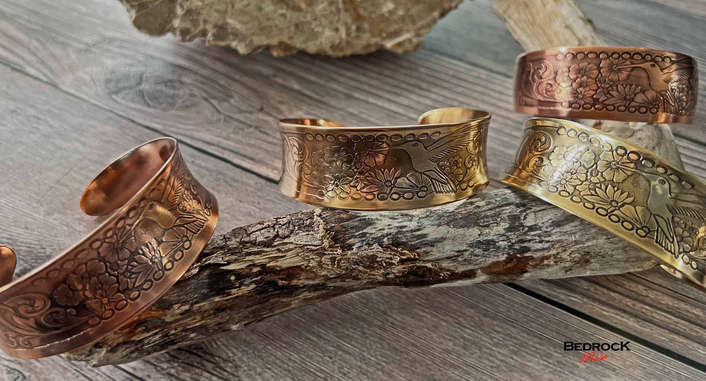 Bedrock Rose hummingbird engraved metal cuffs in copper and brass displayed on a rustic wooden branch.