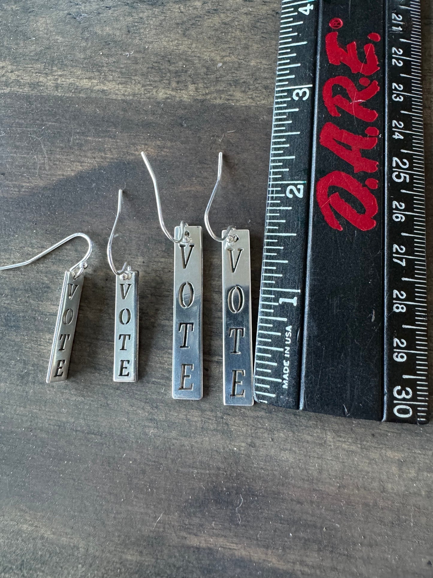 Bedrock Rose silver dangling earrings with the word "Vote" engraved vertically on rectangular pendants. A ruler shows the earrings are approximately 1.5 to 2 inches long.