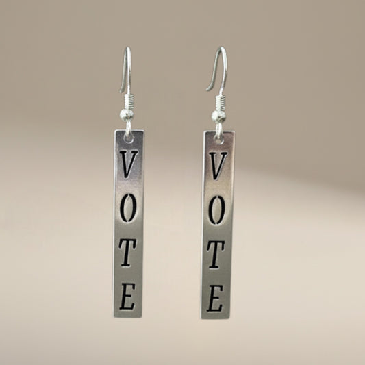 Bedrock Rose silver dangling earrings with the word "Vote" in block letters. Two sizes shown against a dark wood background.