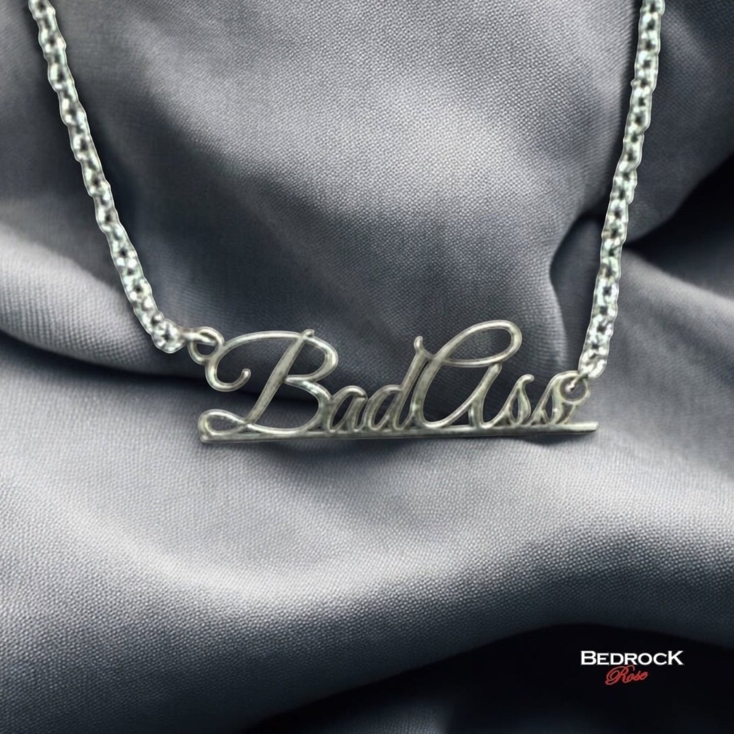 Bedrock Rose sterling silver Bad Ass script pendant necklace on a stainless steel chain against grey fabric
