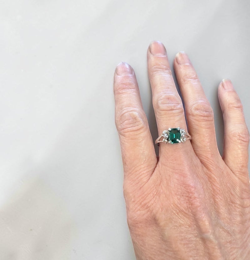 Bedrock Rose emerald cut emerald ring with diamond accents on a silver band, worn on a finger.