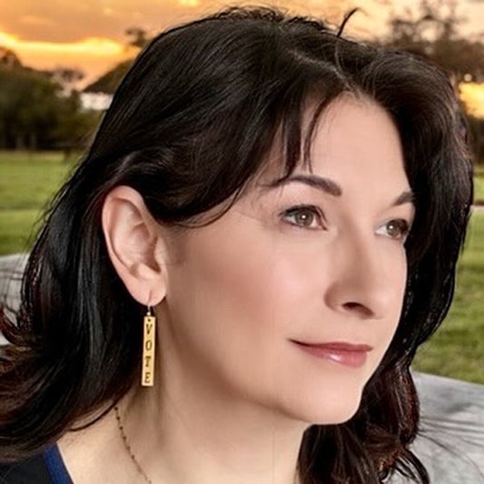 Bedrock Rose gold-plated dangling earrings with laser-cut "Vote" text, worn by a woman outdoors.