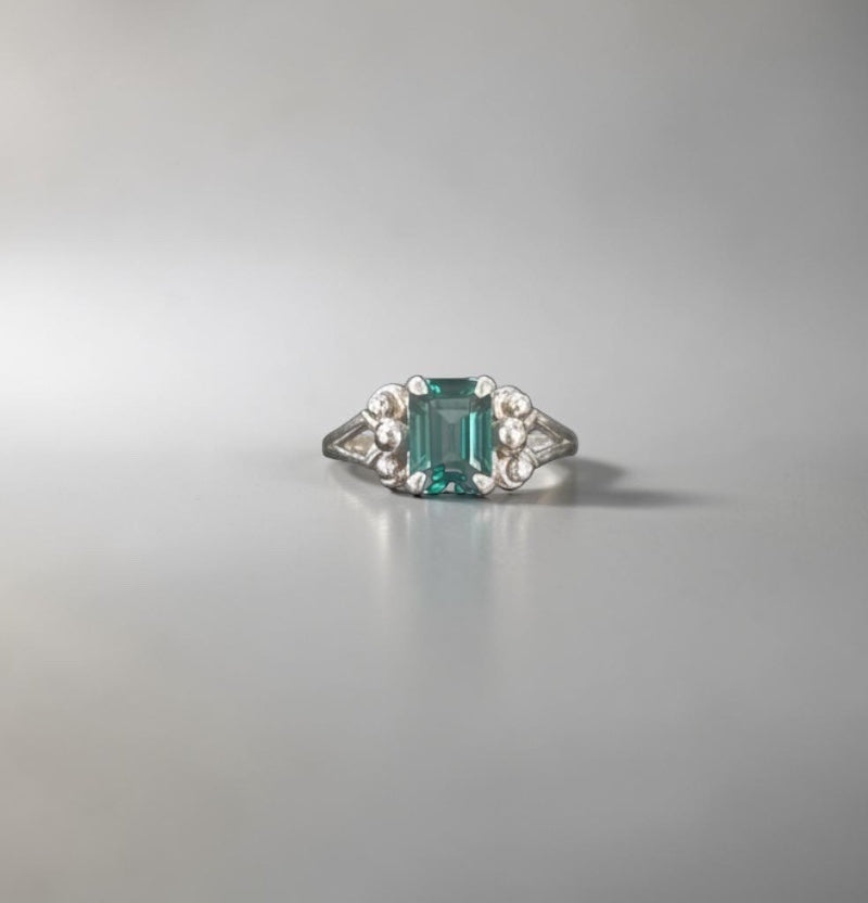 Bedrock Rose emerald-cut teal gemstone ring with silver band and floral accents.